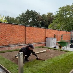 F.H.U Dębowa Kraina - Układanie trawy z rolki w ogrodzie: pracownik wyrównuje podłoże wałkiem, widoczna ceglana ściana, trawa z rolki na obrzeżach, w tle narzędzia ogrodnicze.