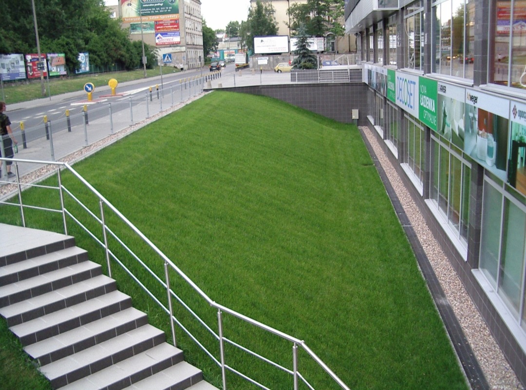 Zadbany trawnik na skarpie przy budynku biurowym, widok z góry ze schodów z metalową barierką. Równa, zielona powierzchnia trawy.