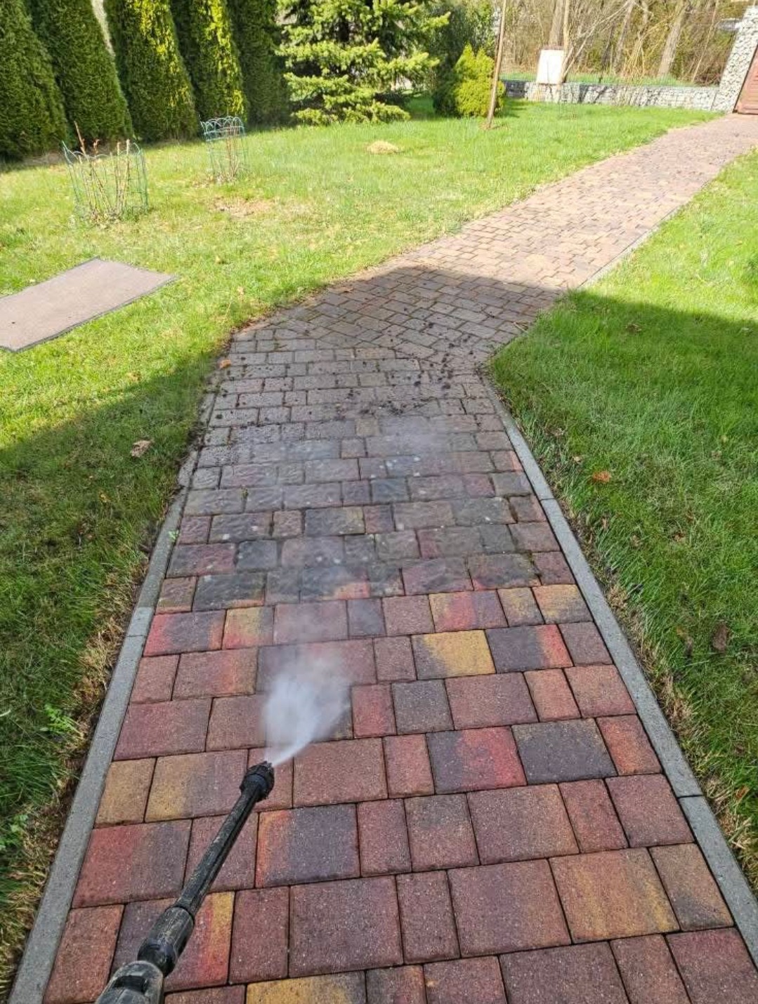 Czyszczenie kostki brukowej myjką ciśnieniową na ścieżce w ogrodzie. Widoczna różnica w kolorze między czystą a brudną powierzchnią. Trawa po bokach ścieżki.