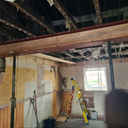 Usługi budowlane - Wnętrze w trakcie remontu: odsłonięty strop, wzmocnienia stalowe, drabina, ściany w stanie surowym. Widoczne narzędzia i elementy konstrukcyjne.