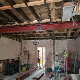 Usługi budowlane - Wnętrze w trakcie przebudowy: stalowa belka podtrzymywana stemplami, odsłonięty strop, widoczne narzędzia i materiały budowlane. Prace przygotowawcze do dalszych etapów.