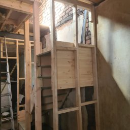 Usługi budowlane - Szkielet schodów z surowego drewna w trakcie budowy, widoczne elementy konstrukcyjne i fragment ściany z cegły. W tle drabina i fragmenty drewnianej konstrukcji.