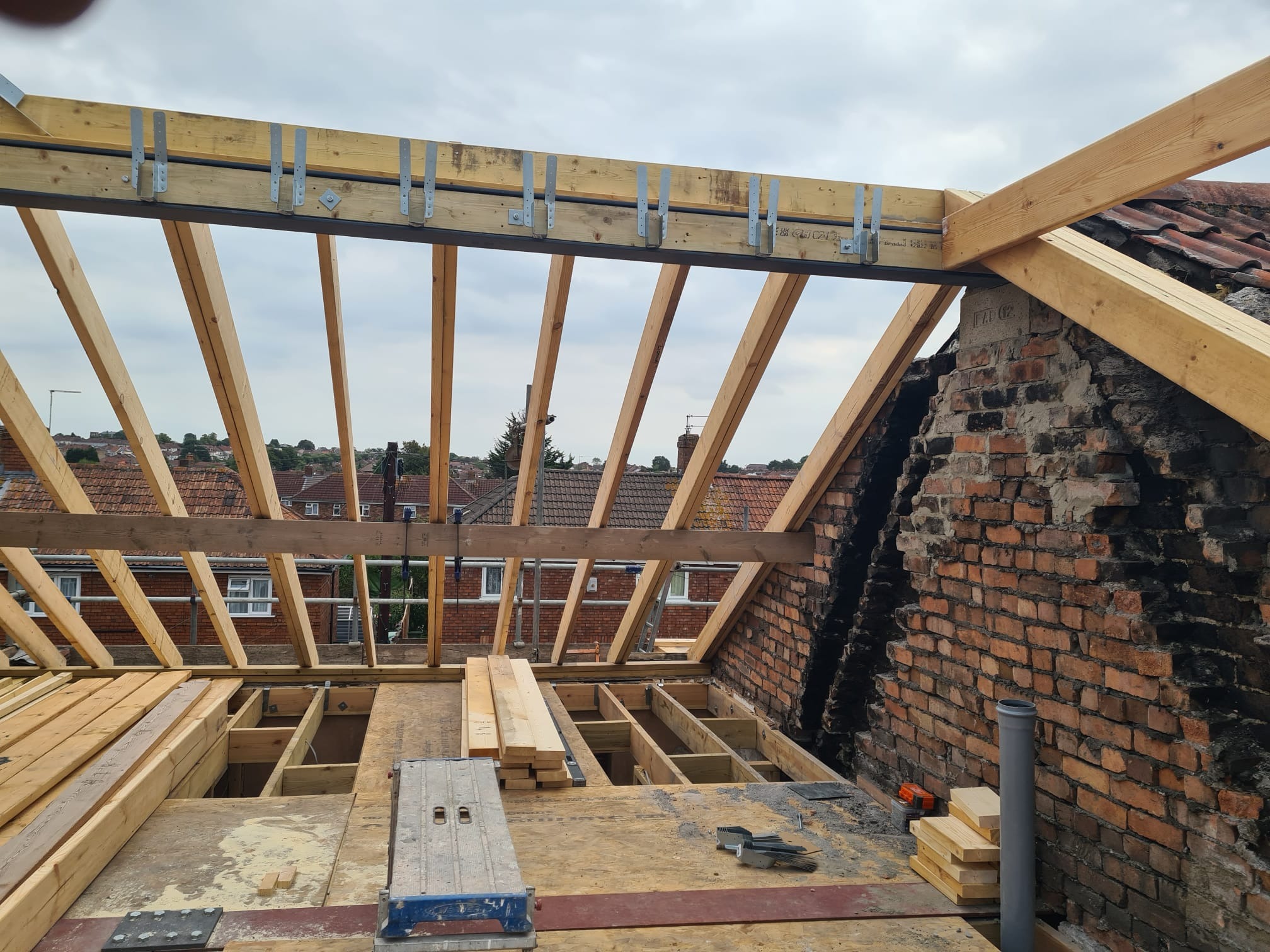 Konstrukcja dachu z drewnianymi belkami i metalowymi łącznikami, widok z góry na budowę z elementami muru z cegły i narzędziami. Etap prac dekarskich.