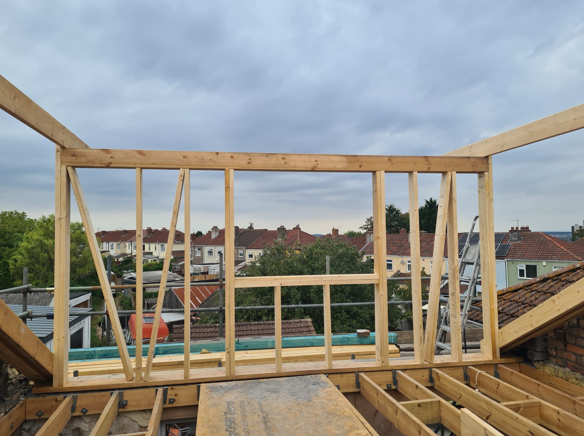 Drewniana konstrukcja szkieletowa na dachu domu, widok na okoliczne budynki i pochmurne niebo. Etap budowy z widocznymi elementami konstrukcyjnymi i rusztowaniem.