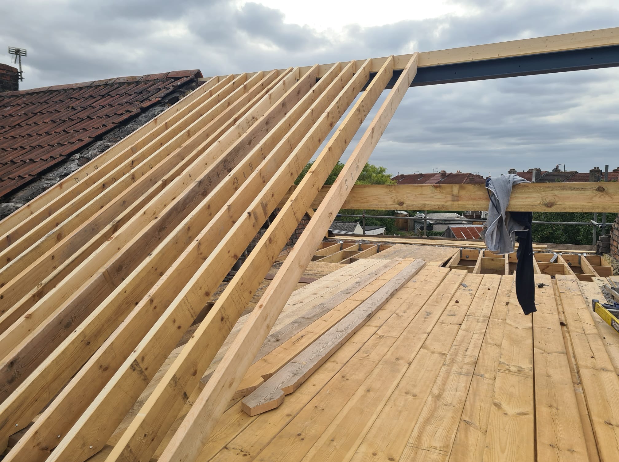 Konstrukcja drewniana dachu w trakcie budowy, widoczne krokwie i deski poszycia, na tle pochmurnego nieba i zabudowań miejskich. Częściowo widoczny stary dach z dachówką.