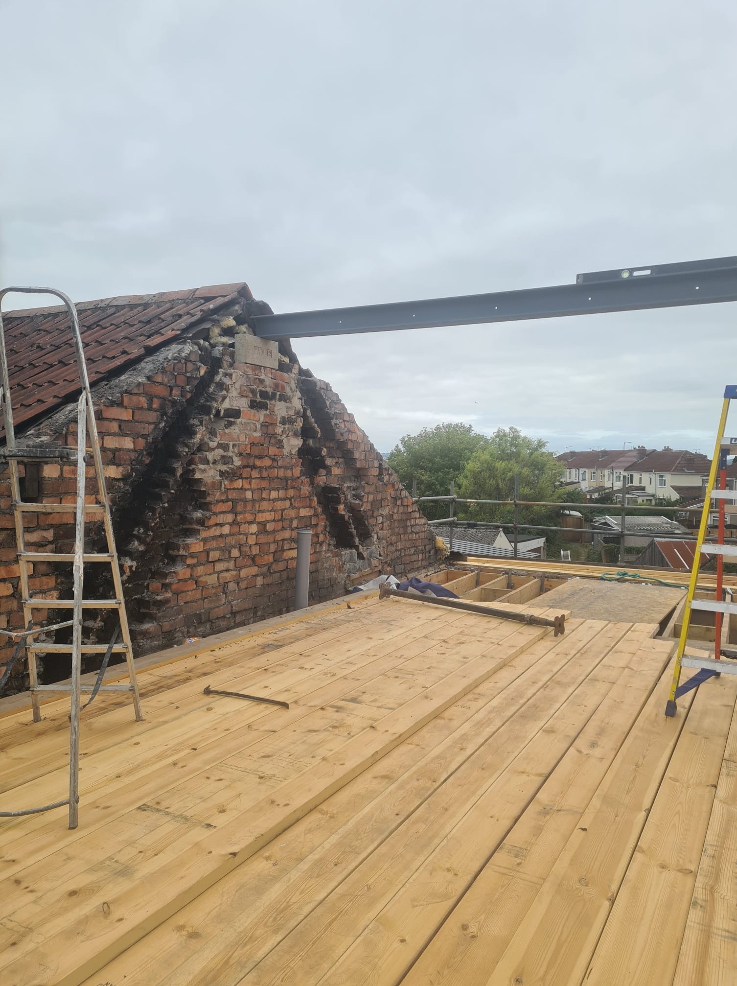 Montaż drewnianej podłogi na dachu budynku z ceglaną ścianą szczytową. Widoczne drabiny i poziomica na stalowej belce. Prace budowlane w toku.