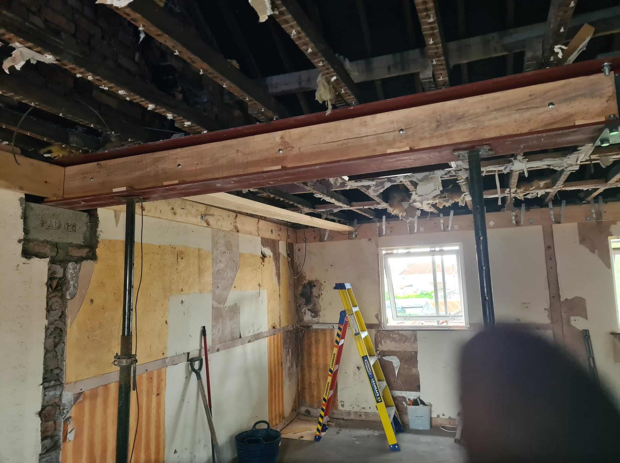 Wnętrze w trakcie remontu: odsłonięty strop, wzmocnienia stalowe, drabina, ściany w stanie surowym. Widoczne narzędzia i elementy konstrukcyjne.