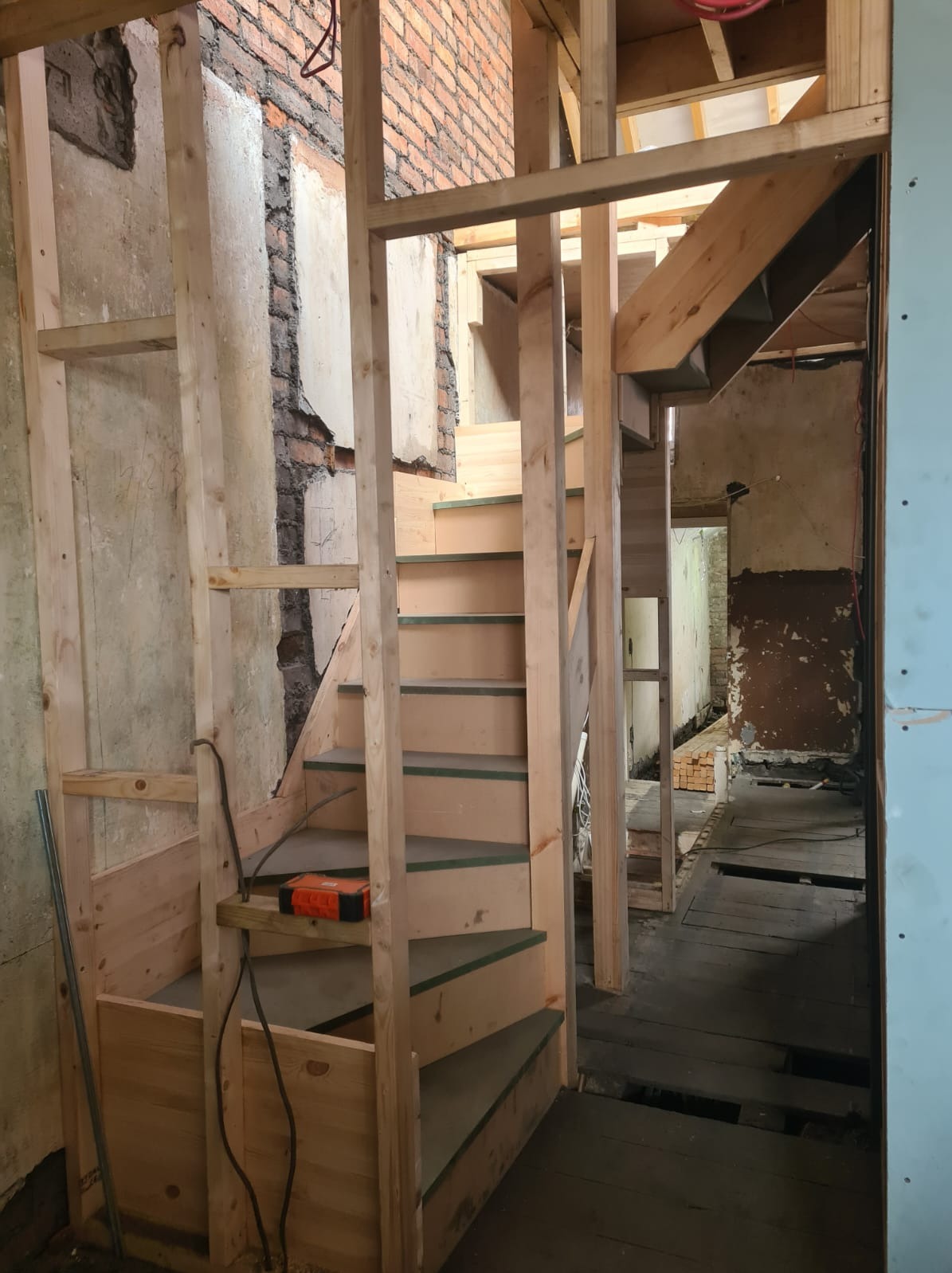 Wnętrze w trakcie remontu: drewniana konstrukcja schodów na tle surowych ścian z odsłoniętą cegłą i fragmentami tynku. Widoczne elementy instalacji i narzędzia.