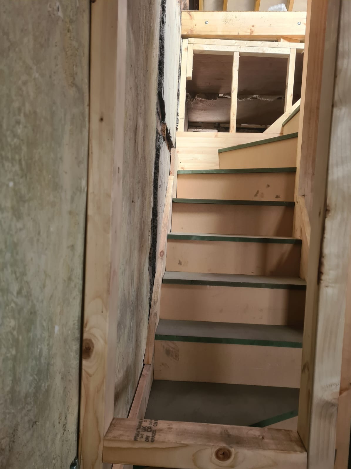 Wnętrze w trakcie prac: drewniane schody z zielonymi akcentami, surowe ściany i konstrukcja stropu. Widok z dołu, ukazujący etap budowy lub przebudowy.