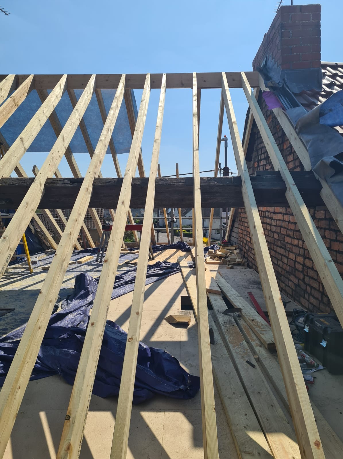 Konstrukcja drewniana dachu w trakcie budowy, widok z dołu na niebo. Elementy więźby dachowej, ceglany komin w tle, niebieska plandeka zabezpieczająca.