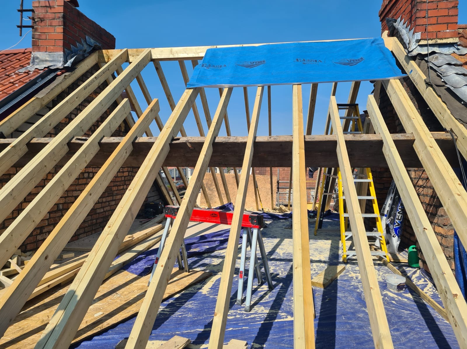 Konstrukcja dachu z drewnianych belek, zabezpieczona niebieską membraną, widok z dołu na tle błękitnego nieba. Widoczne narzędzia i drabina. Etap budowy dachu.