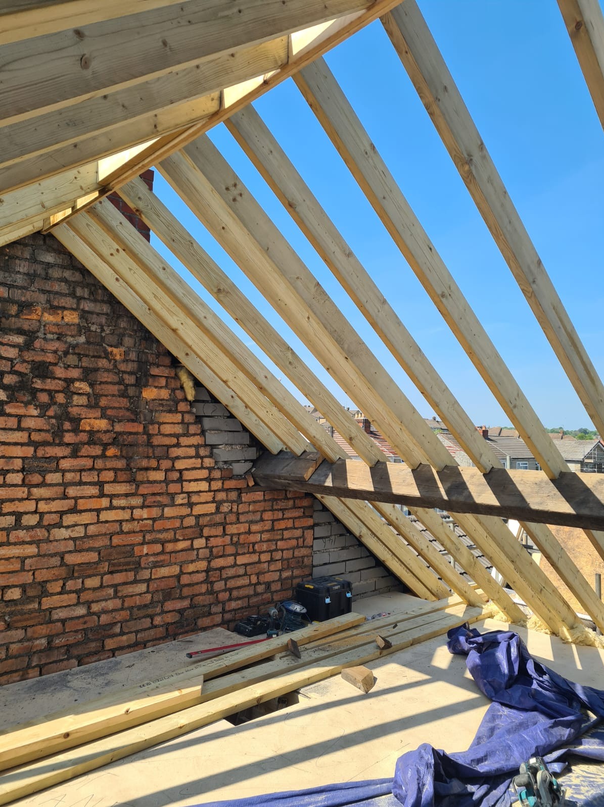 Konstrukcja drewniana dachu w trakcie budowy, widok z wnętrza. Czerwona cegła ściany szczytowej i narzędzia budowlane w tle. Błękitne niebo widoczne przez krokwie.