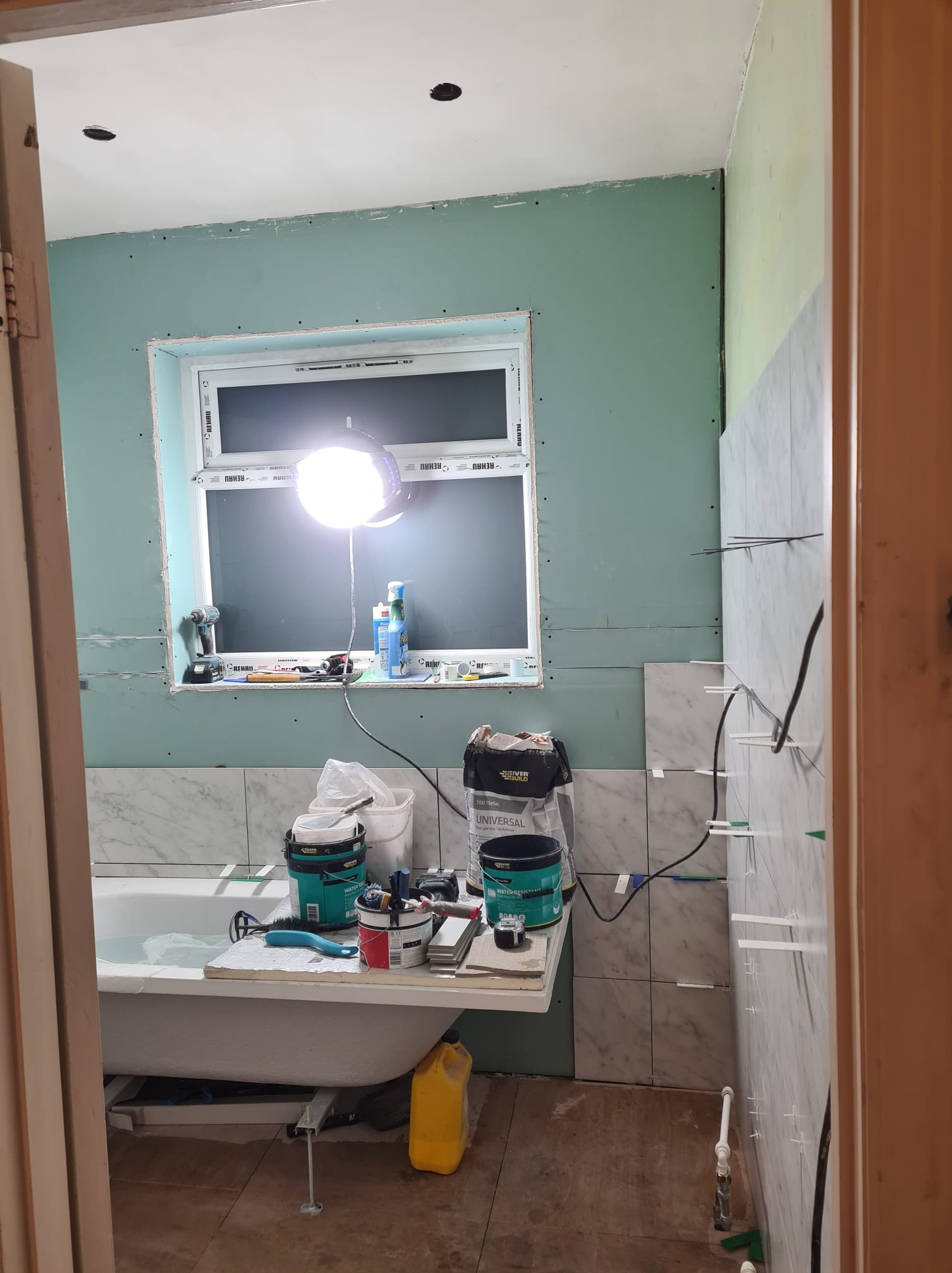 Łazienka w trakcie remontu: widoczna wanna, okno z narzędziami, ściany z płyt gipsowych i częściowo ułożone marmurowe płytki. Oświetlenie robocze.