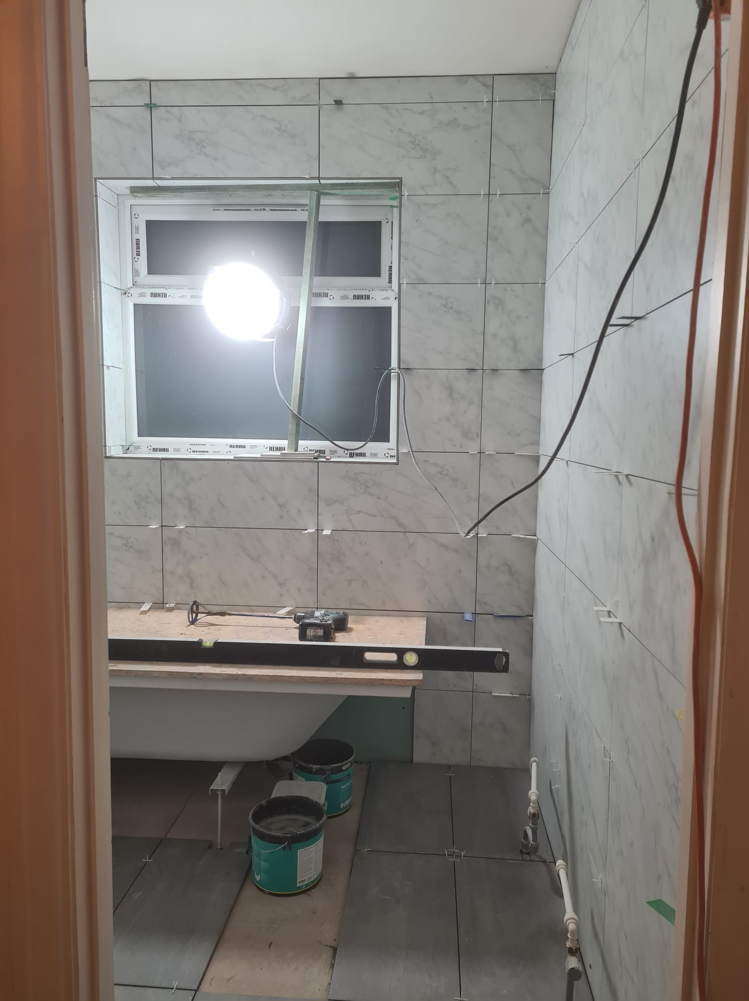 Remont łazienki w trakcie: białe płytki imitujące marmur na ścianach, szare płytki na podłodze, wanna, narzędzia i lampa budowlana. Widok zza futryny.