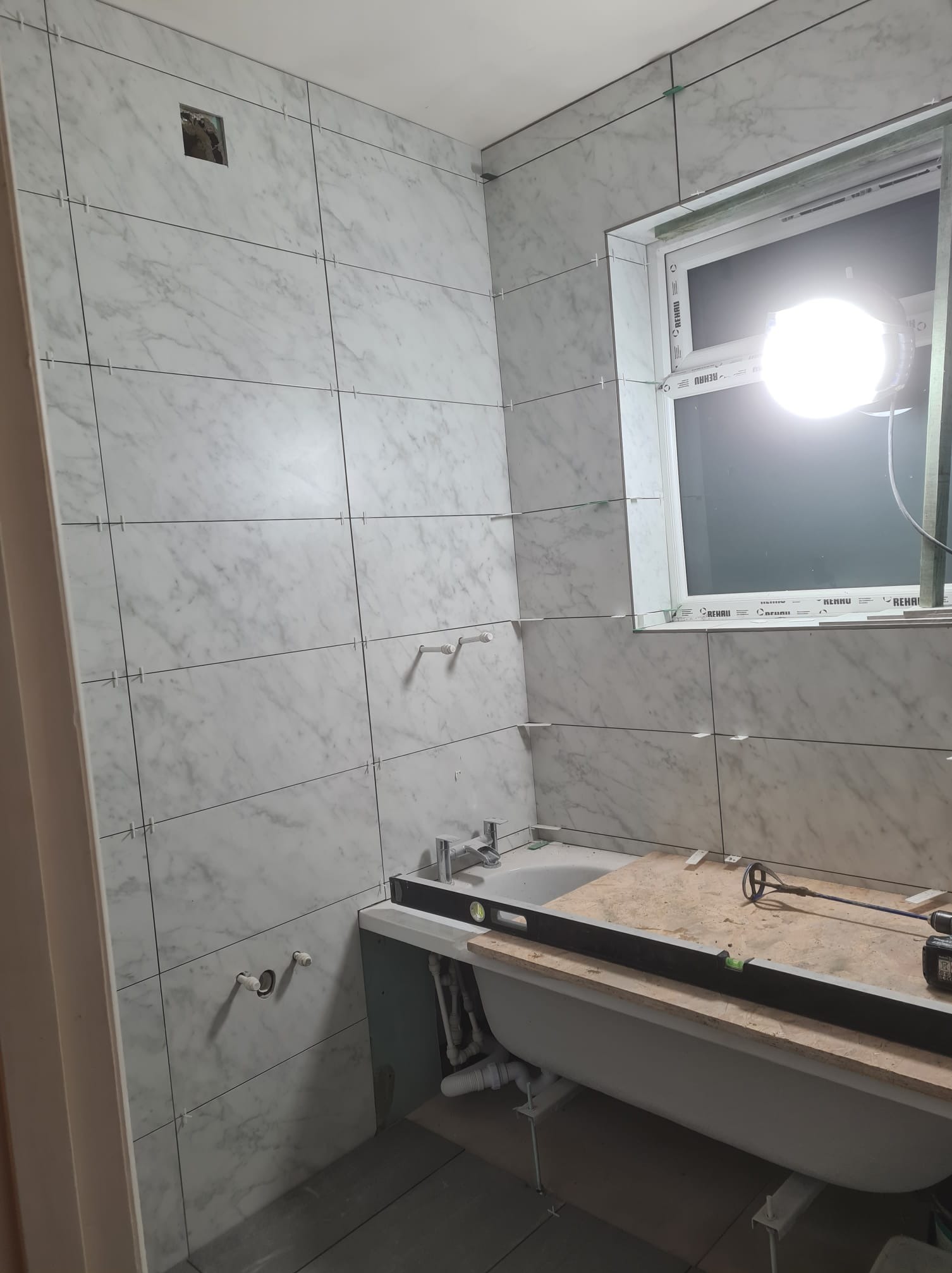 Białe marmurowe płytki ścienne w trakcie montażu w łazience, widoczne krzyżyki dystansowe, poziomica na obudowie wanny i okno z jasnym światłem.