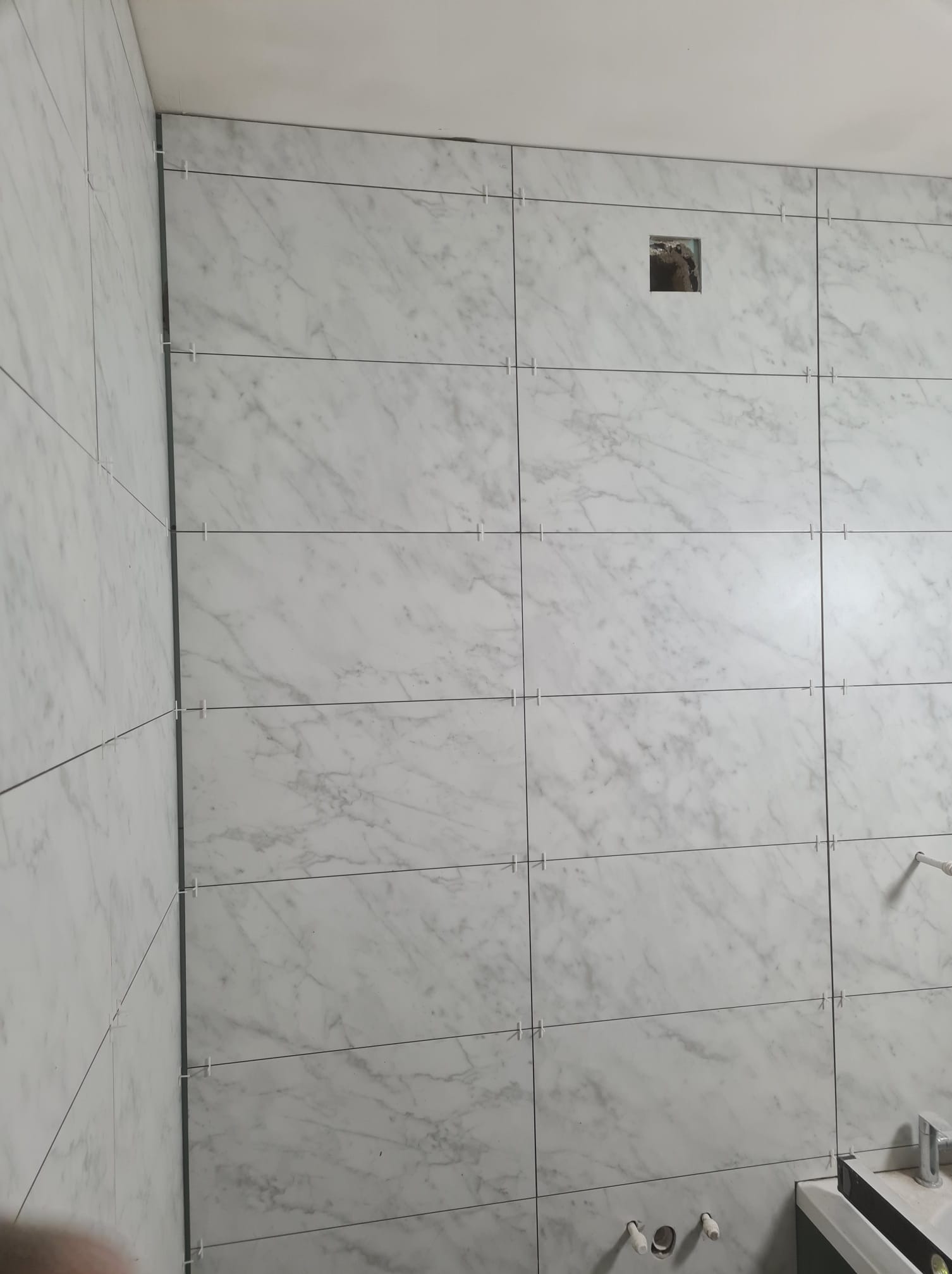 Ściana w trakcie układania płytek imitujących marmur, z krzyżykami dystansowymi, widoczny otwór na instalację. Ujęcie z bliska, z perspektywy osoby stojącej.