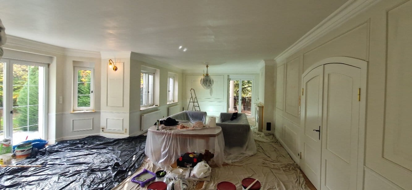 Wnętrze pokoju z białymi ścianami w trakcie prac przygotowawczych do malowania. Podłoga i meble zabezpieczone folią, widoczne narzędzia i farby.