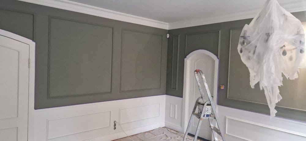 Pomieszczenie po malowaniu z dekoracyjnymi panelami ściennymi w odcieniach szarości i bieli. Drabina malarska i zabezpieczony żyrandol wskazują na świeżo zakończone prace.