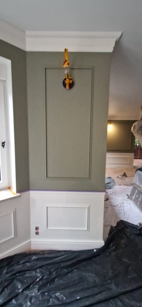 Ściana po malowaniu z dekoracyjnymi listwami w kolorze oliwkowym i białym. Na podłodze folia ochronna. Widoczne narzędzia malarskie w tle.