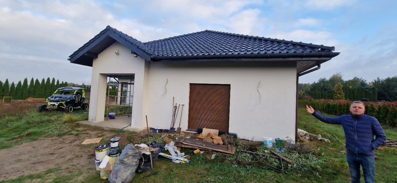 Otynkowany budynek gospodarczy z ciemną dachówką, z widocznymi śladami prac wykończeniowych i osobą wskazującą na elewację. W tle zieleń i pojazd terenowy.