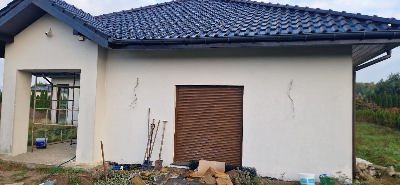 Świeżo otynkowana elewacja domu z brązową roletą i narzędziami malarskimi. Widoczne rusztowania i wiaderka z farbą. Tło stanowi zieleń ogrodu.