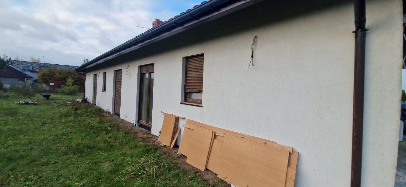 Boczny widok domu z jasną elewacją, brązowymi oknami i roletami. Na trawie leżą płyty. Widoczne ślady po instalacji na ścianie.