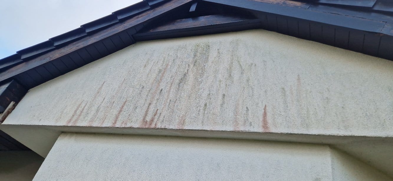 Zabrudzona elewacja budynku z widocznymi zaciekami i śladami organicznymi pod okapem dachu. Widoczne ciemne drewniane elementy konstrukcji dachu.