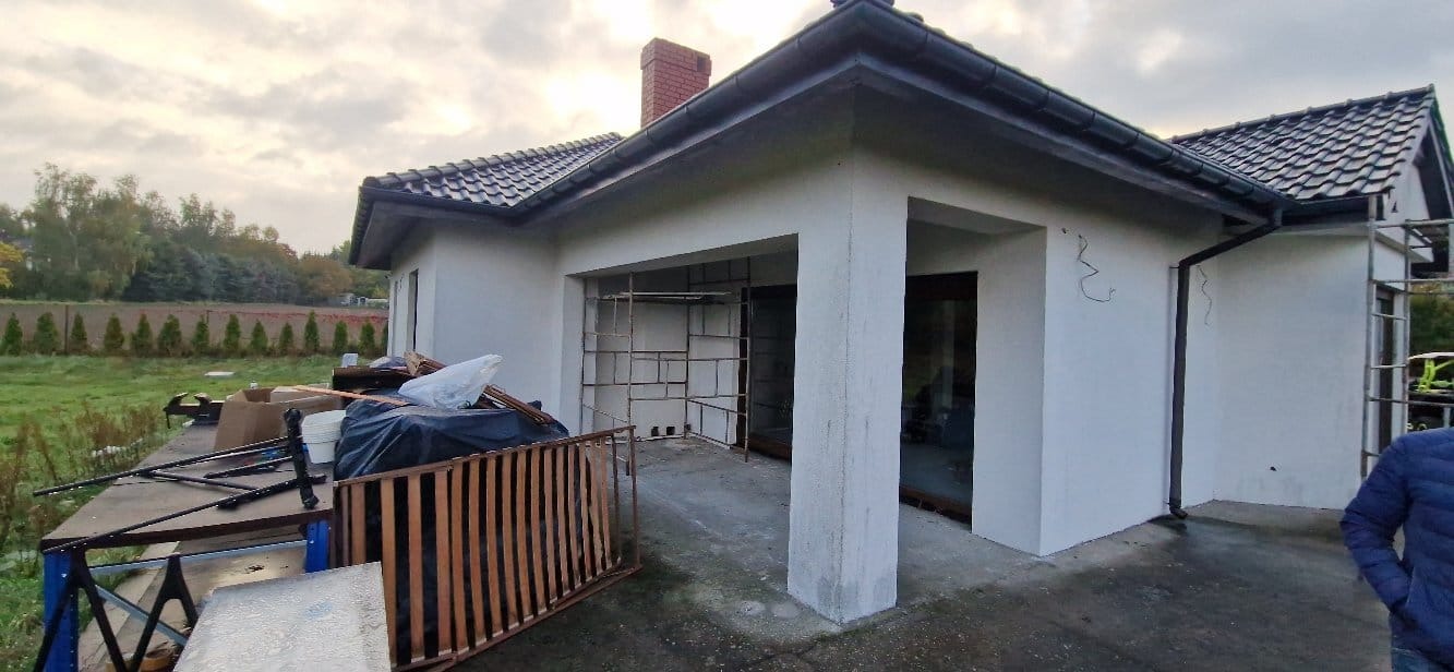 Biała elewacja domu jednorodzinnego po nałożeniu gładzi, z widocznymi rusztowaniami i elementami przygotowania do dalszych prac wykończeniowych.