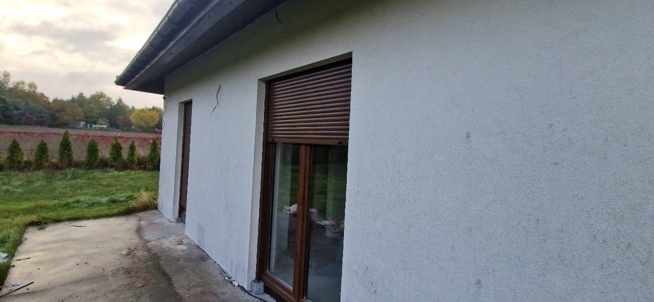 Elewacja budynku po nałożeniu szarego tynku strukturalnego, widoczne okna z brązowymi roletami zewnętrznymi i pęknięcia na ścianie. Tło: trawnik i drzewa.