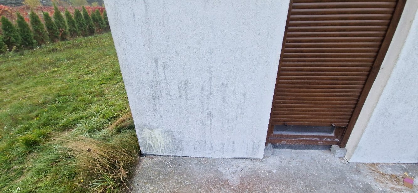 Fragment elewacji z brudnymi zaciekami przy brązowej rolecie okiennej. Widoczne trawy i tuje w tle. Niska jakość wykonania malowania.