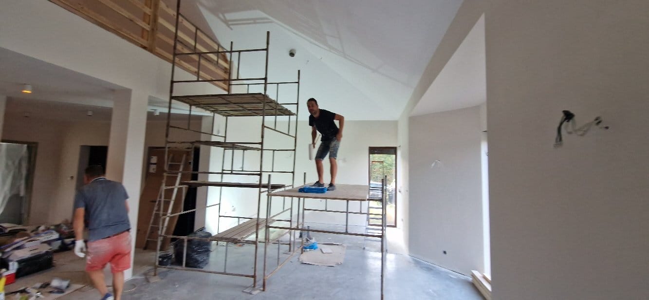 Wnętrze w trakcie malowania: mężczyzna na rusztowaniu z wałkiem, świeżo pomalowane ściany, widoczne kable. Drugi mężczyzna w oddali. Praca w toku.