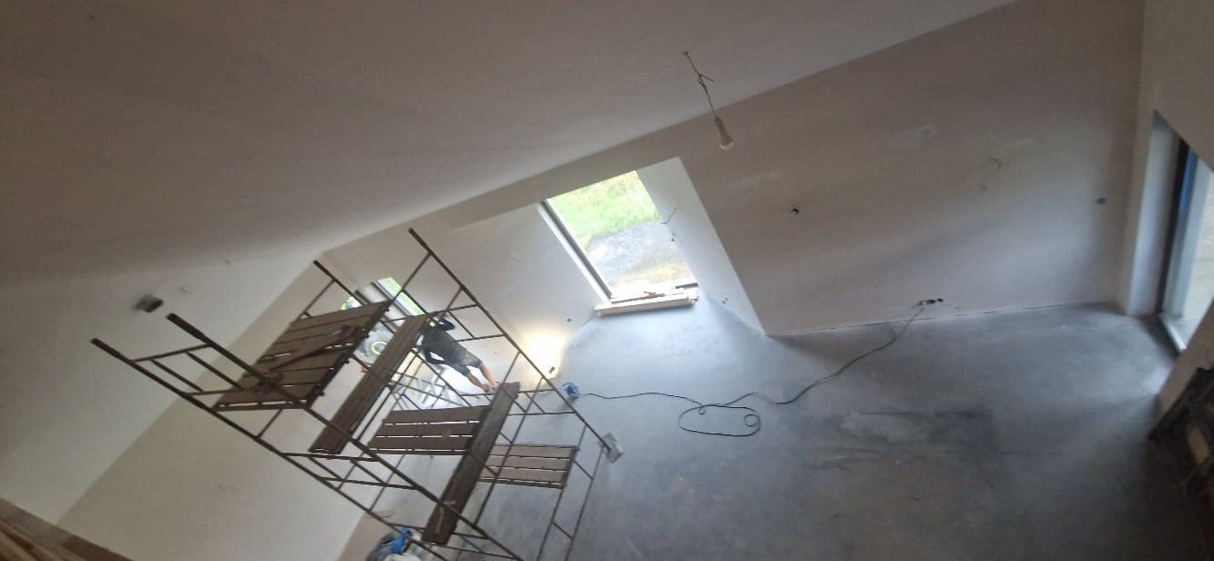 Skośne poddasze po szpachlowaniu, widok z góry. Na pierwszym planie rusztowanie, w tle okno dachowe i ściana z oknem. Widoczne ślady po kablach elektrycznych.
