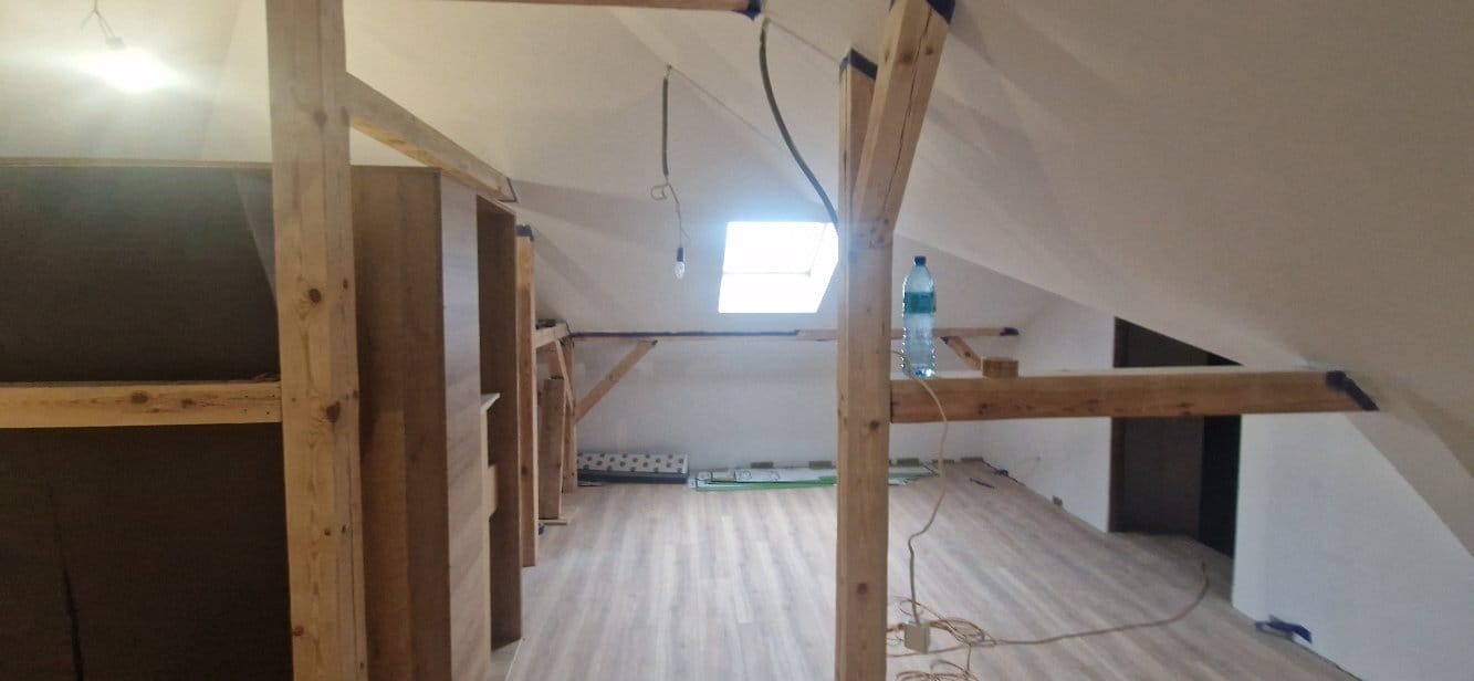 Poddasze ze skośnymi ścianami po remoncie. Jasne ściany, drewniane elementy konstrukcyjne i panele podłogowe. Widoczne okno dachowe i butelka wody.