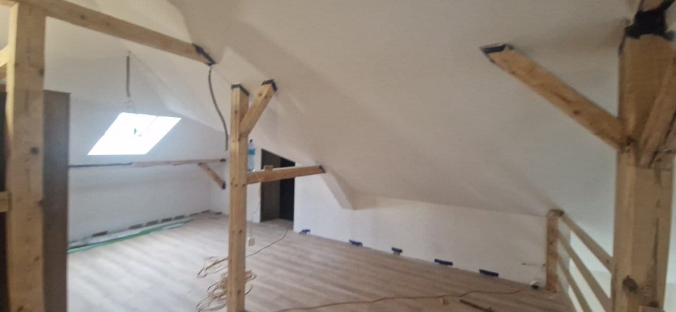 Białe ściany poddasza z drewnianą konstrukcją nośną i oknem dachowym. Widoczne ślady po taśmie malarskiej na podłodze. Świeżo pomalowane wnętrze.