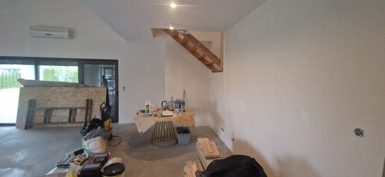 Wnętrze w trakcie prac wykończeniowych: gładkie, białe ściany, widoczne narzędzia i materiały budowlane, fragment drewnianej konstrukcji schodów. Przygotowanie do malowania.