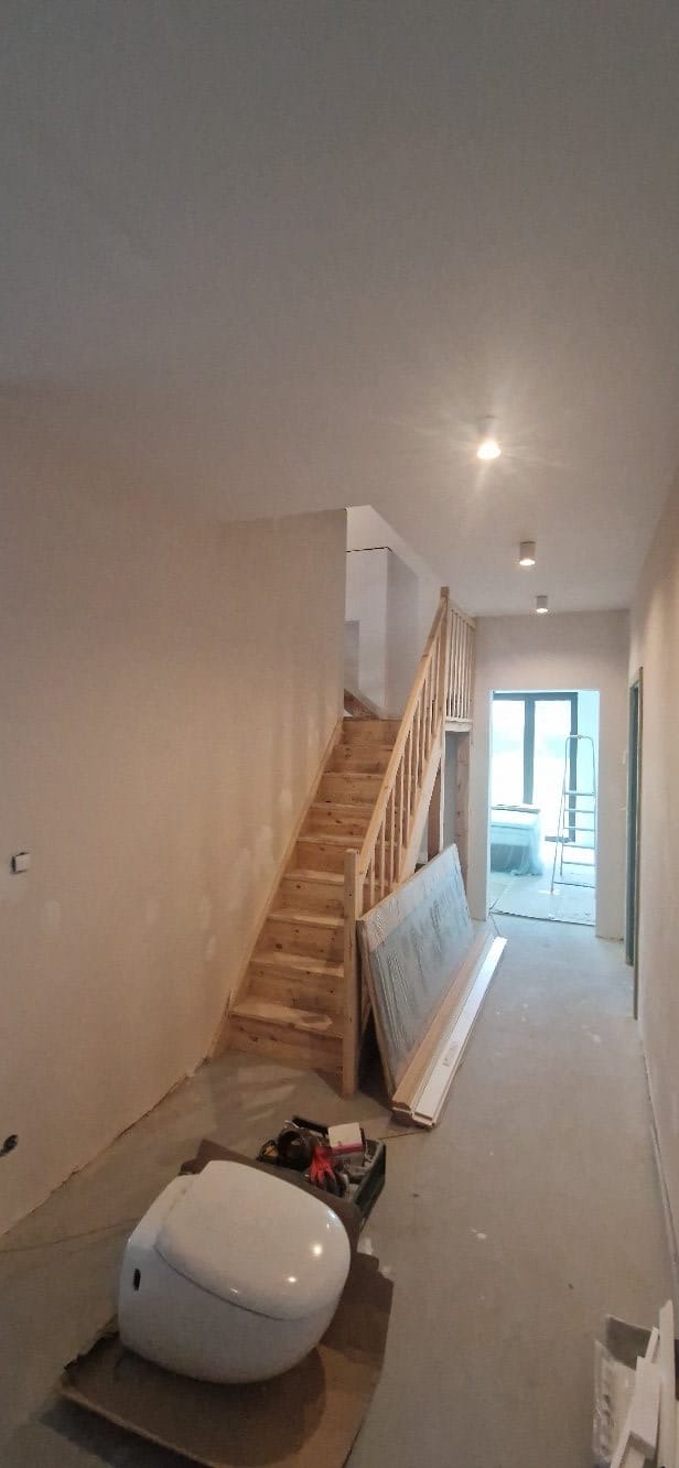 Wnętrze w trakcie szpachlowania: surowe ściany, drewniane schody, narzędzia i sanitariat na podłodze, przygotowane do dalszych prac wykończeniowych.