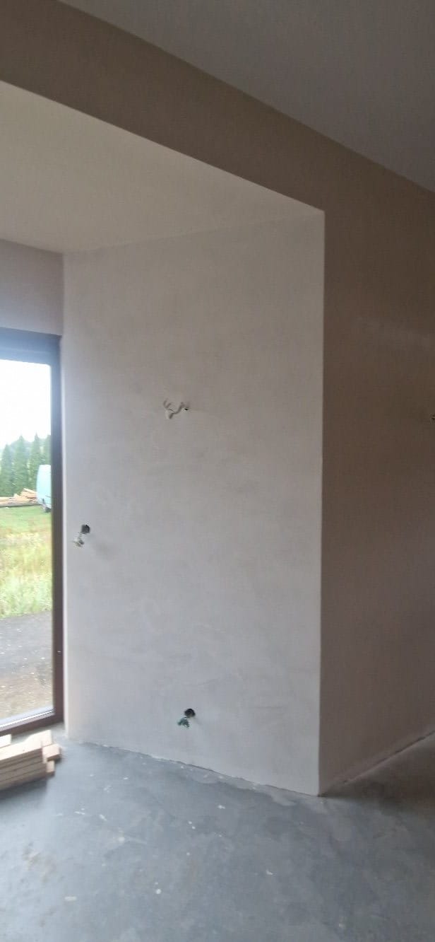 Wnętrze w trakcie prac wykończeniowych: gładka, jasna ściana z widocznymi punktami elektrycznymi, surowa podłoga, okno z widokiem na zewnątrz. Przy oknie leżą deski.