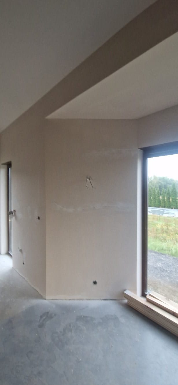 Wnętrze pokoju po szpachlowaniu ścian, widoczne gładkie powierzchnie przygotowane do malowania. Okno z widokiem na zewnątrz. Podłoga betonowa.