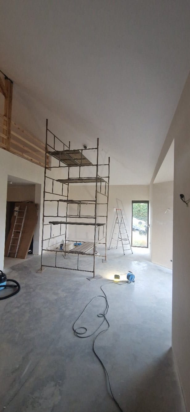 Wnętrze w trakcie prac wykończeniowych: ściany po szpachlowaniu, widoczne rusztowanie, drabina i sprzęt budowlany. Przygotowanie do malowania w nowym domu.