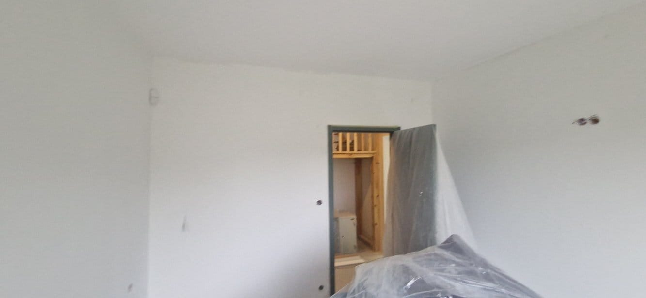 Wnętrze po świeżym malowaniu na biało. Widoczne gładkie ściany, zabezpieczone meble folią i otwór drzwiowy prowadzący do kolejnego pomieszczenia.