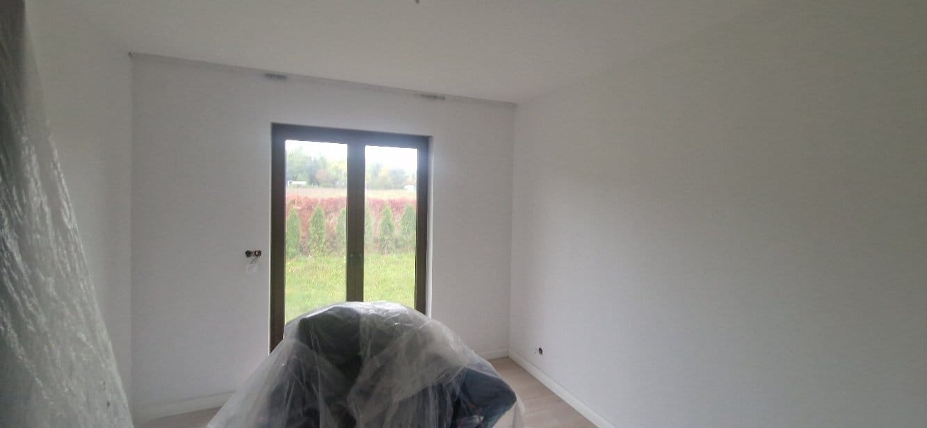 Świeżo pomalowane, białe ściany w pokoju z oknem w Dąbrówce. Krzesło zabezpieczone folią malarską. Minimalistyczne wykończenie wnętrza po remoncie.