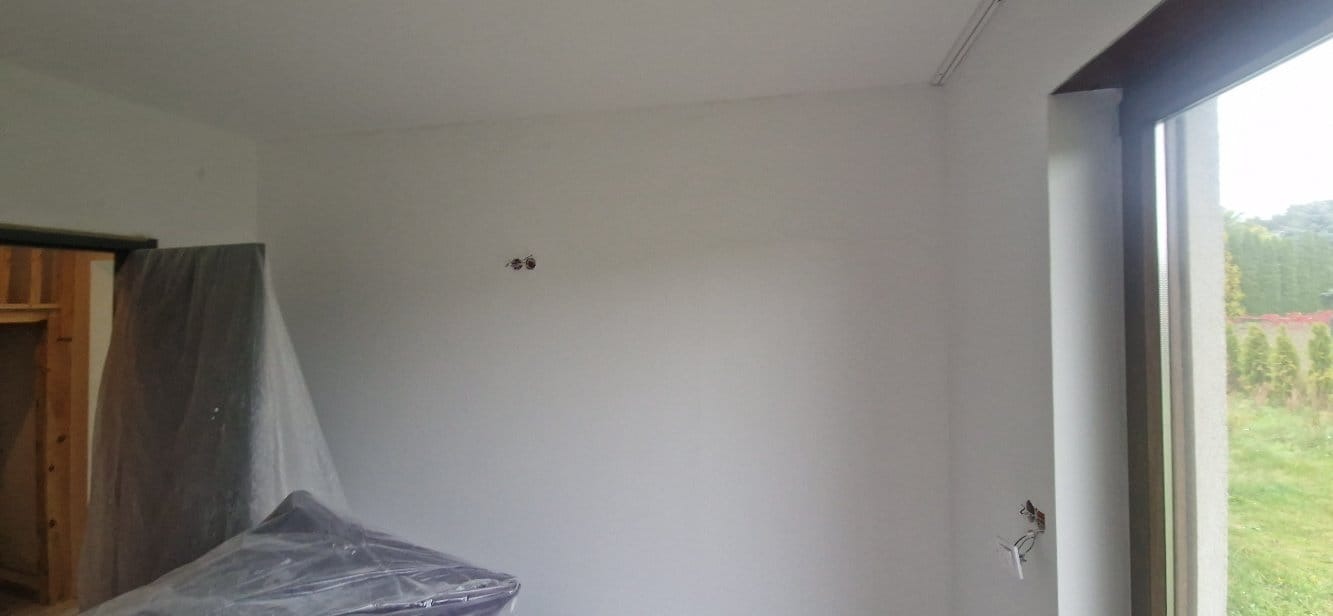 Wnętrze pokoju po szpachlowaniu ścian, przygotowane do malowania. Widoczne gładkie ściany, zabezpieczone meble folią i wystające kable elektryczne.