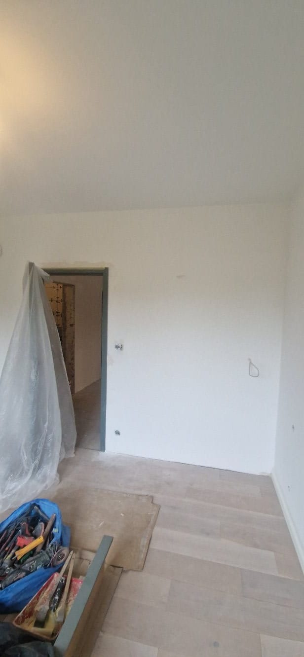 Wnętrze pokoju po malowaniu ścian na biało, z widocznymi narzędziami malarskimi i zabezpieczonym wejściem. Podłoga wyłożona panelami w odcieniach beżu.