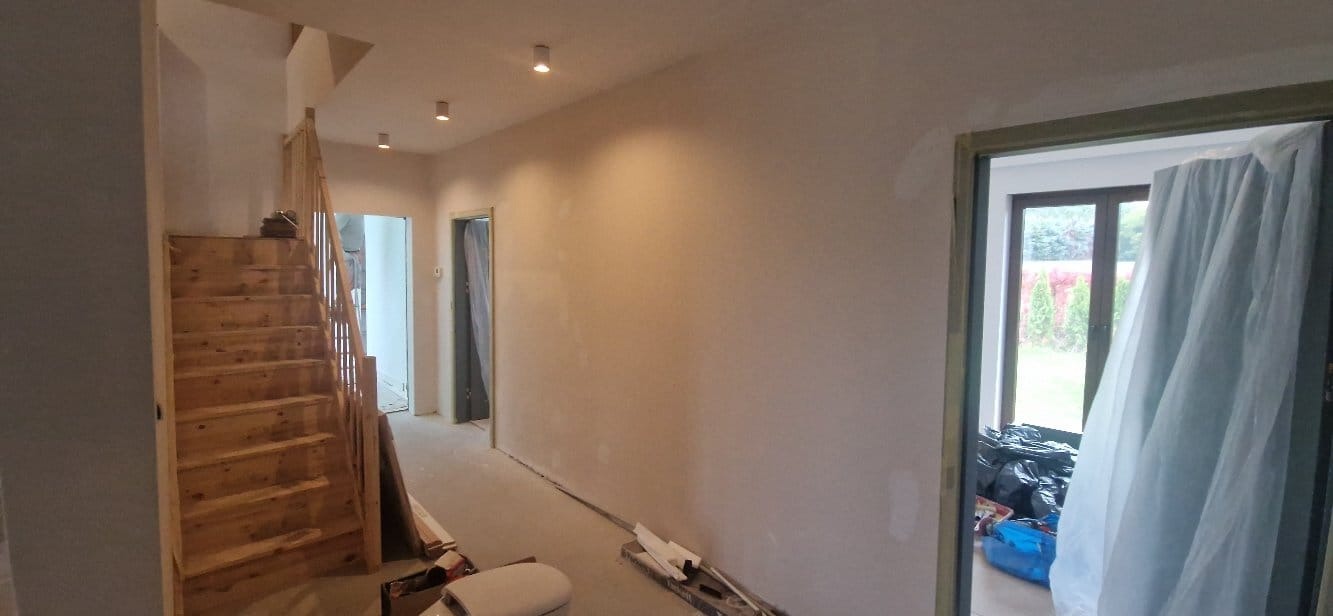 Wnętrze domu po szpachlowaniu ścian, widoczne drewniane schody i przygotowane do malowania pomieszczenia z oknem na ogród.