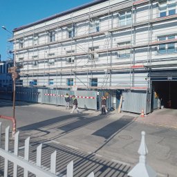 Krystian Wróbel BELBUD - Budynek w rusztowaniach, zabezpieczony metalowymi panelami, z pracownikami w odblaskowych kamizelkach. Widok zza ozdobnego, białego ogrodzenia.