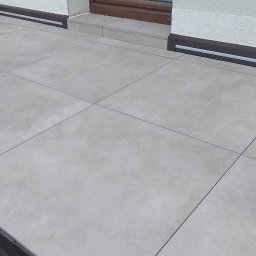 Krystian Wróbel BELBUD - Jasnoszara, kwadratowa podłoga z płytek na zewnątrz budynku, z widocznym oknem i fragmentem ściany. Widoczne fugi i listwy przypodłogowe.