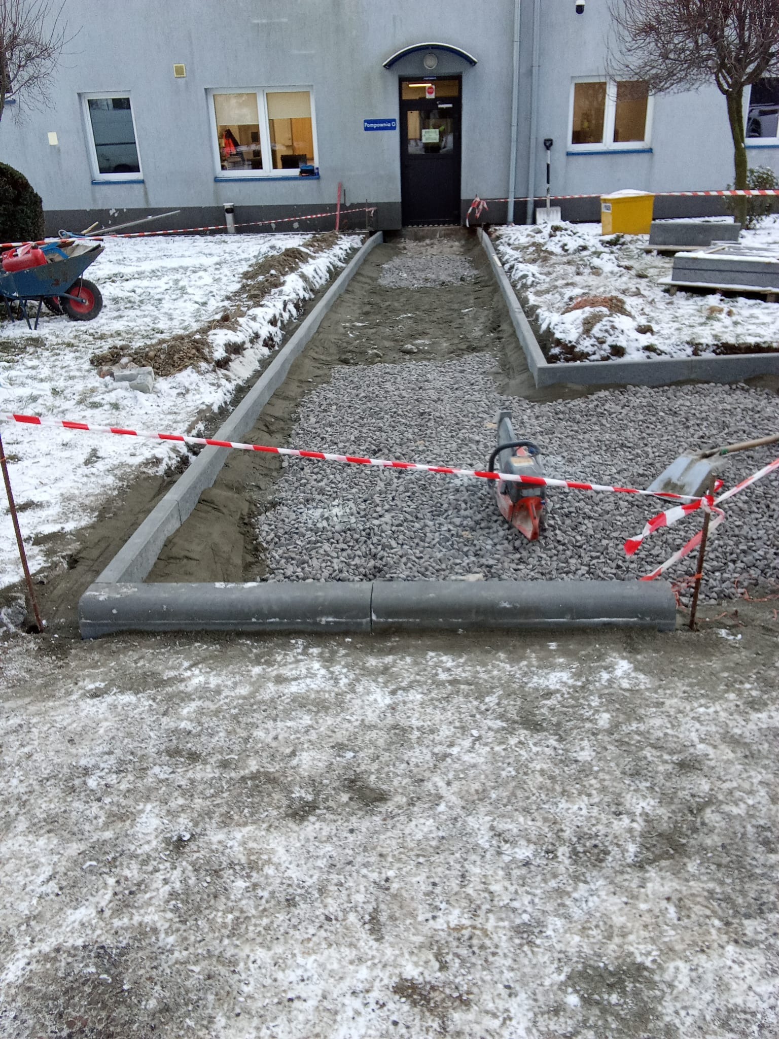 Układanie chodnika z kostki brukowej przed budynkiem w zimowej scenerii. Widoczna ubijarka, szpadel i taśma ostrzegawcza.