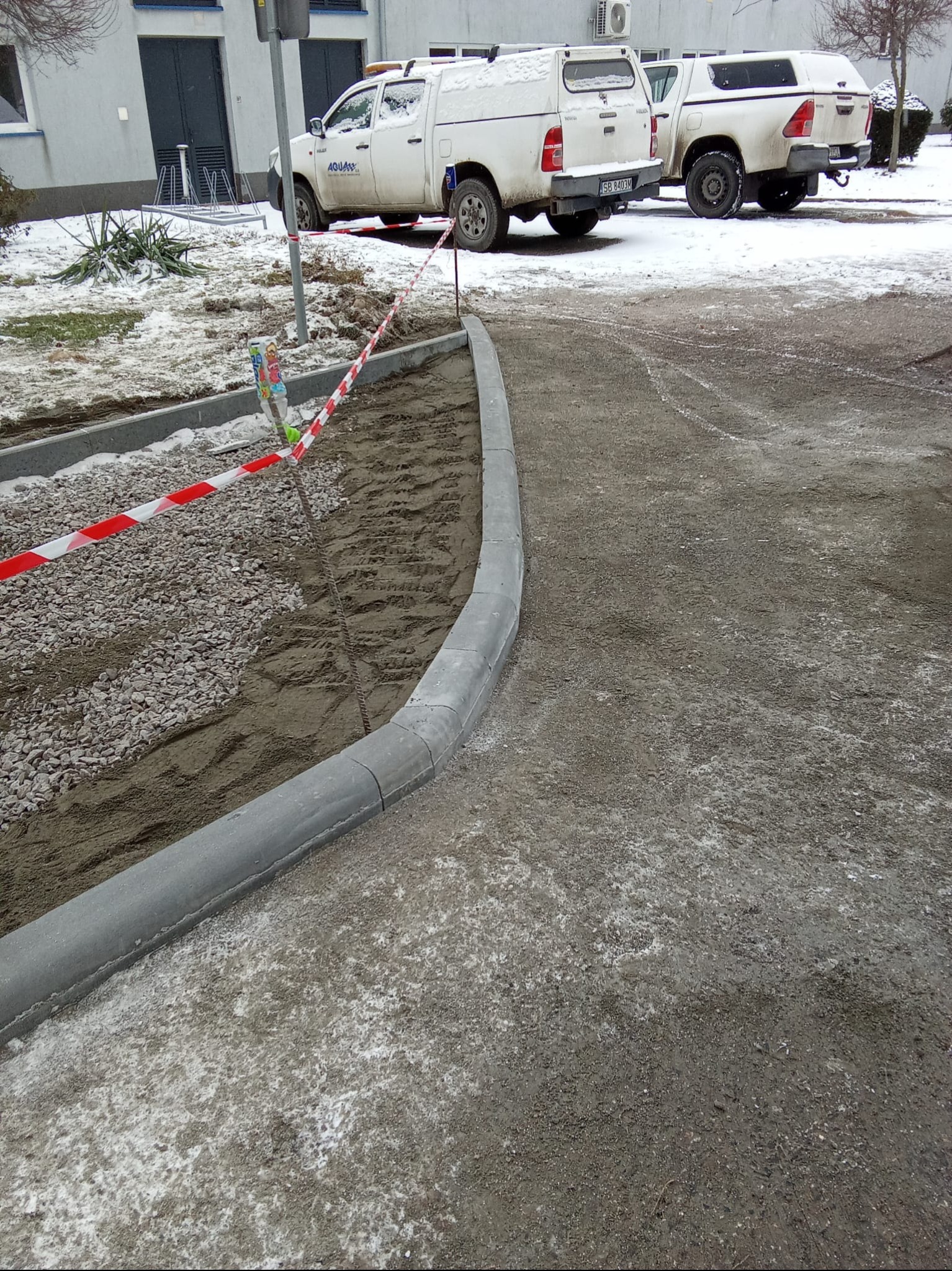 Układanie obrzeży chodnikowych w Sosnowicach. Widoczne przygotowane podłoże, obrzeża oraz teren zabezpieczony taśmą. W tle zaparkowane samochody.