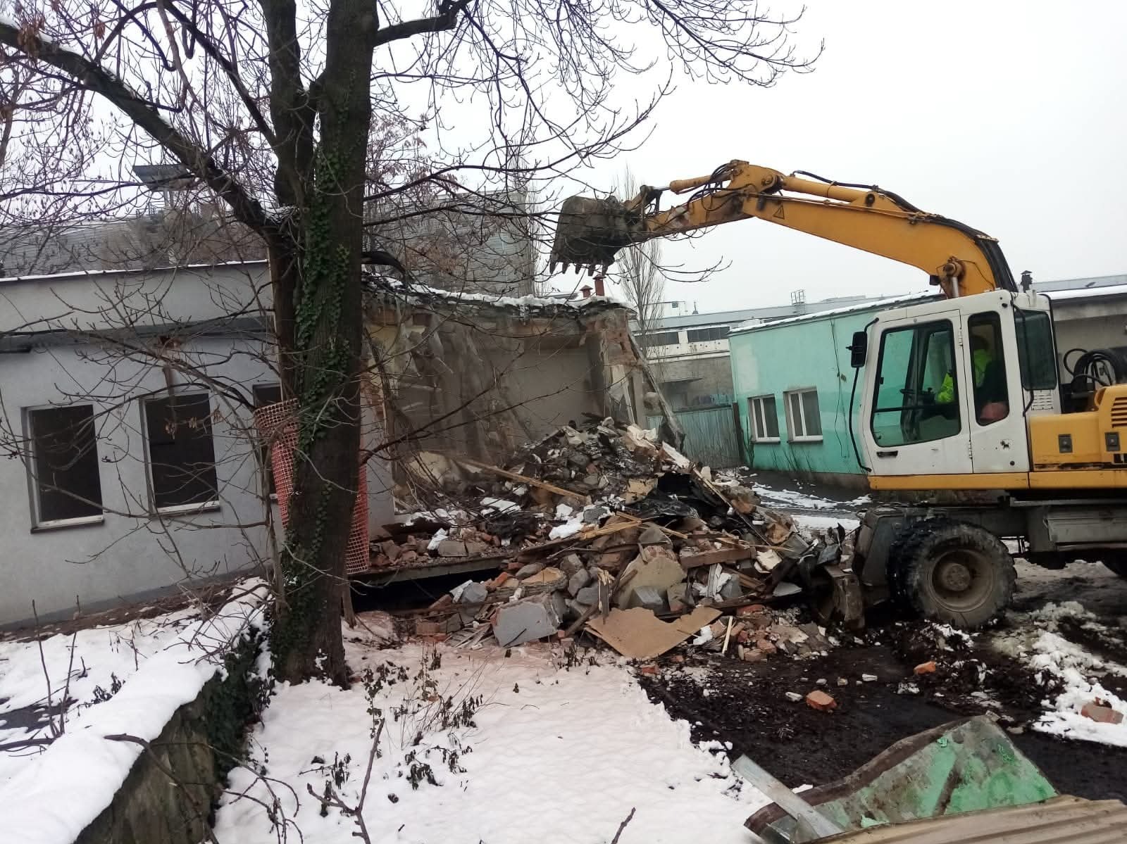 Żółta koparka kołowa w trakcie rozbiórki budynku, z widocznymi gruzami i fragmentami konstrukcji. W tle budynek z zieloną elewacją i drzewo bez liści.