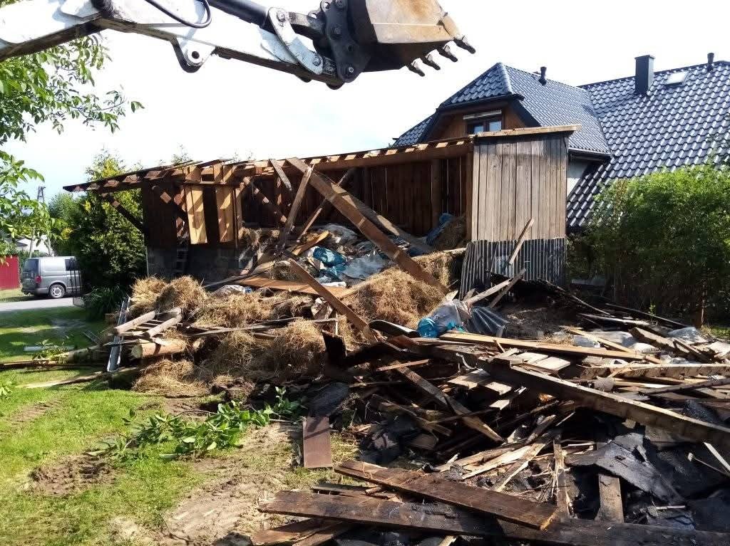 Demontaż drewnianej konstrukcji przez koparkę na tle domu z ciemną dachówką. Widoczne pozostałości po rozbiórce i fragment koparki.